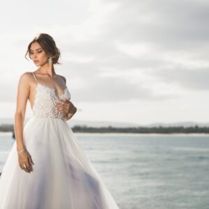 A beautiful bride in a stylish wedding dress posing by the ocean under soft daylight.